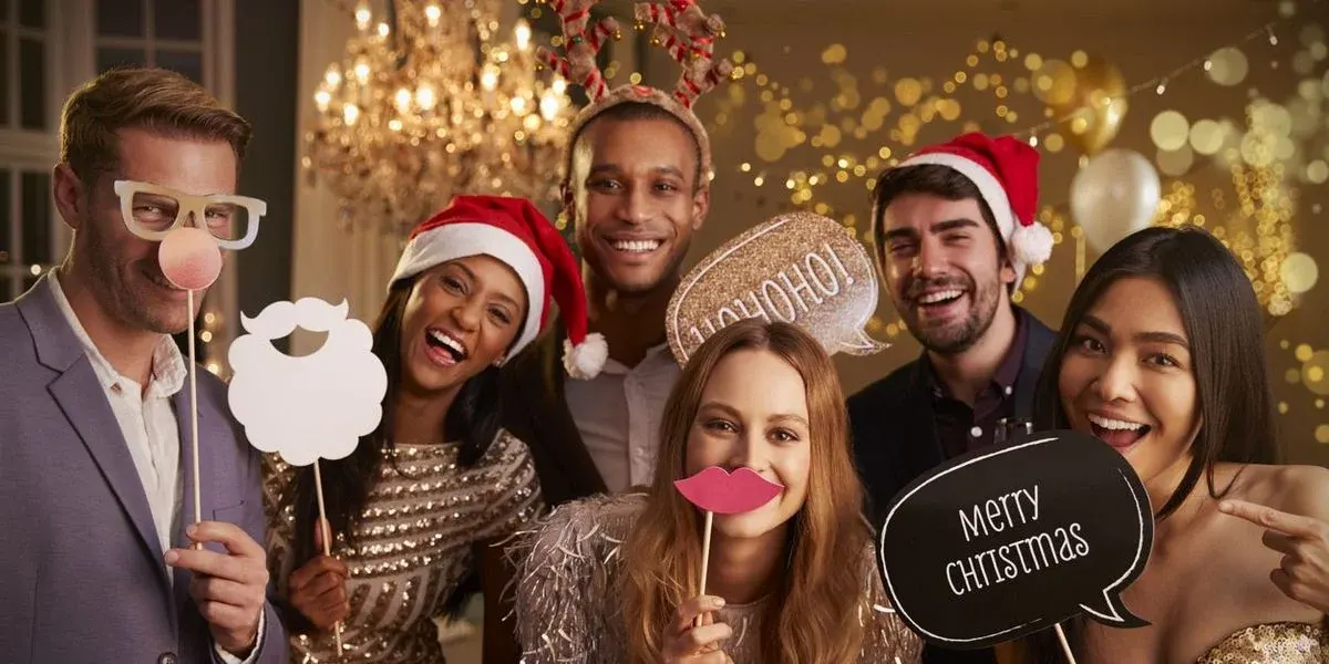 Sechs lächelnde Personen halten festliche Fotorequisiten: Weihnachtsmann-Hüte, Rentiergeweih, Bart, Brille, Lippen und Sprechblasen mit "HO HO HO!" und "Merry Christmas".