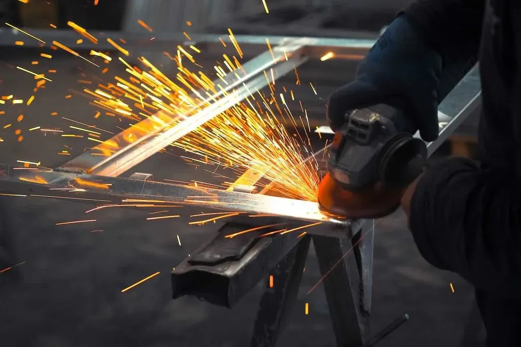 Handwerker mit Schutzhandschuhen bearbeitet Stahlstab mit Winkelschleifer, intensive Funkenspritzer sichtbar