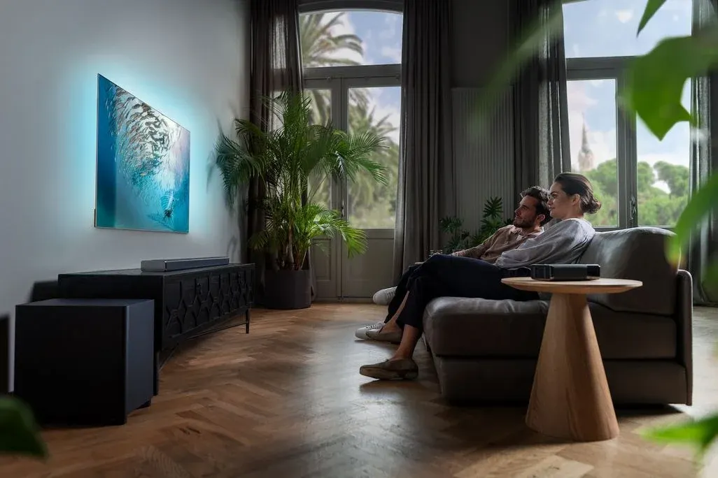 Paar schaut TV mit blauer Unterwasserszene in Wohnzimmer. Davor Soundbar, Subwoofer. Große Fenster, Pflanzen und Bäume im Hintergrund.