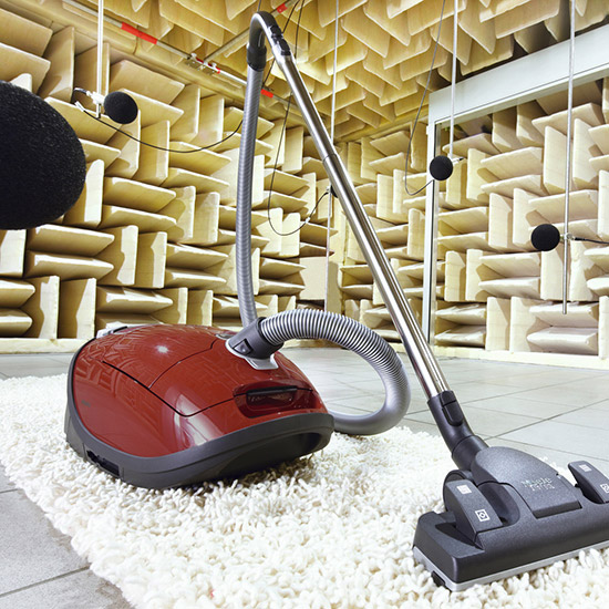 Ein roter Staubsauger steht auf einem Teppich in einem schalltoten Raum mit beigen Pyramidenwänden. Mikrofone hängen von der Decke.