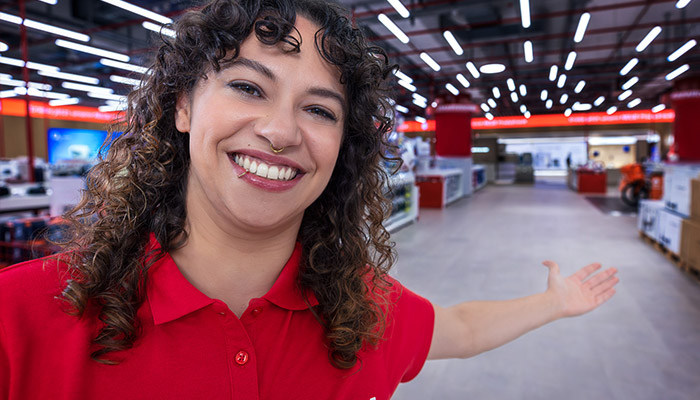 Eine lächelnde Person in einem roten Hemd steht in einem Geschäft mit Regalen und Beleuchtung.