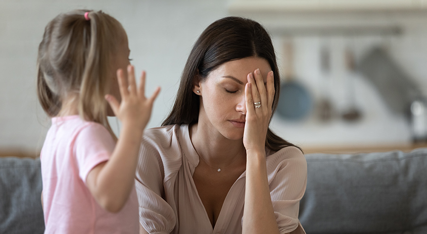 Eine Frau sitzt mit geschlossenen Augen, eine Hand bedeckt ihr Gesicht. Ein Kind steht daneben und hebt eine Hand.