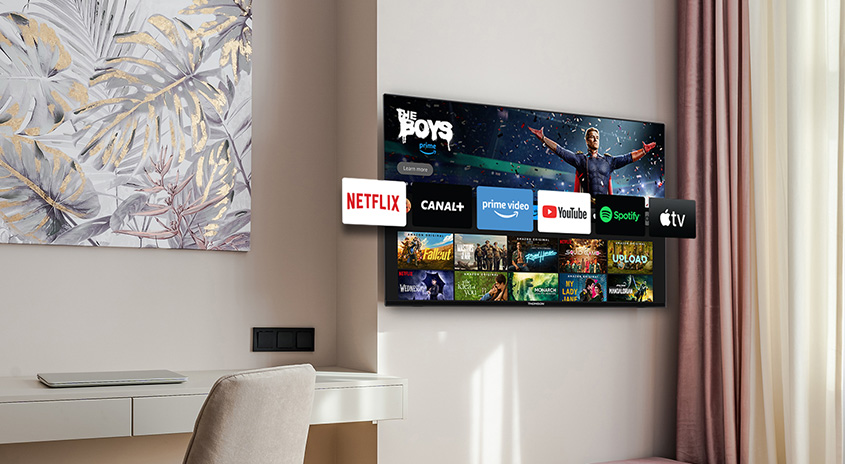 An der Wand hängt ein Fernseher mit Streaming-Apps. Darunter befindet sich ein Schreibtisch mit einem Laptop und einem Stuhl.