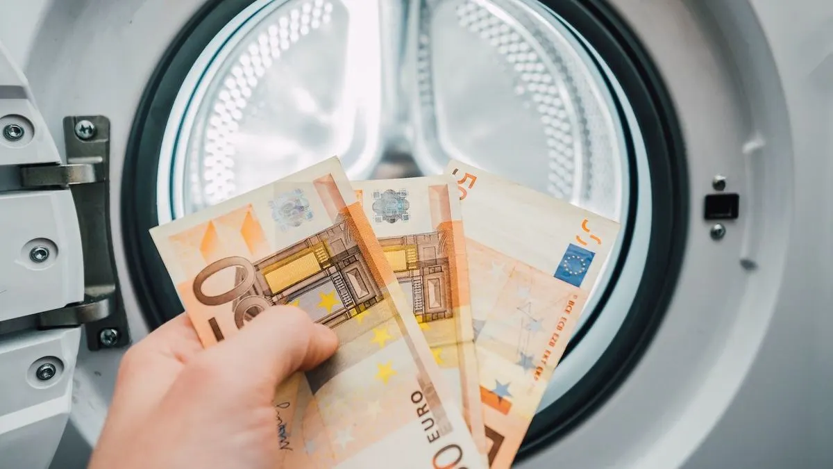 Hand haelt Euro-Geldscheine vor geöffneter Waschmaschinentrommel.