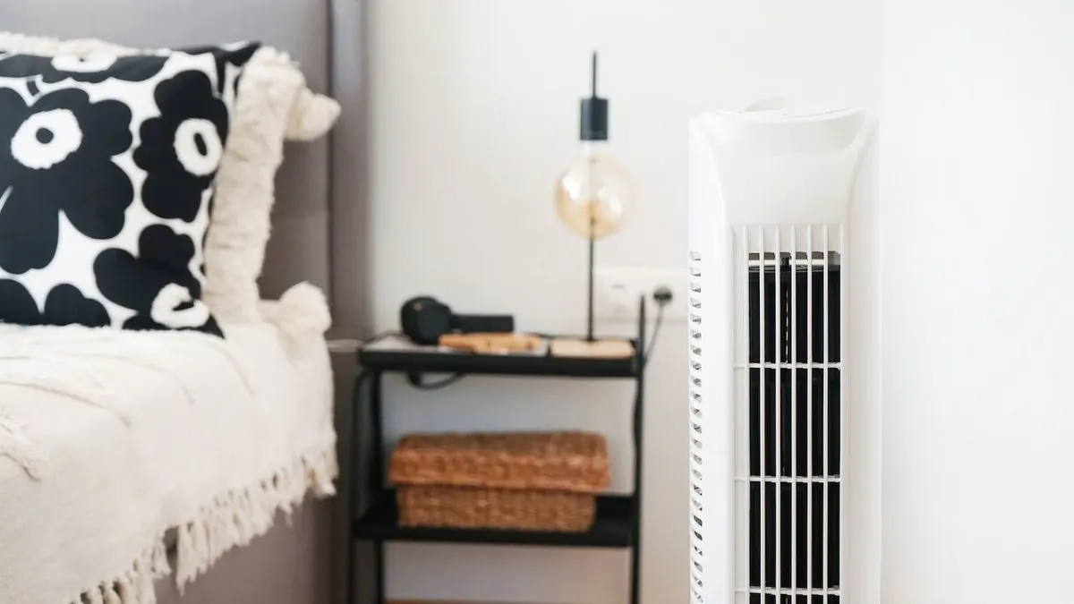 Weißer Turmventilator rechts. Links ein Bett mit schwarz-weißem Kissen, beiger Decke und Nachttisch mit Lampe, Körben.