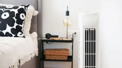 Weißer Turmventilator rechts. Links ein Bett mit schwarz-weißem Kissen, beiger Decke und Nachttisch mit Lampe, Körben.
