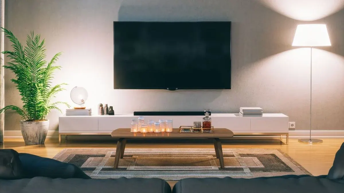 Wohnzimmer mit großem Fernseher an grauer Wand, darunter weiße TV-Bank. Links Grünpflanze, rechts Stehlampe. Couchtisch mit Kerzen und Dekanter auf Teppich vor Sofa.