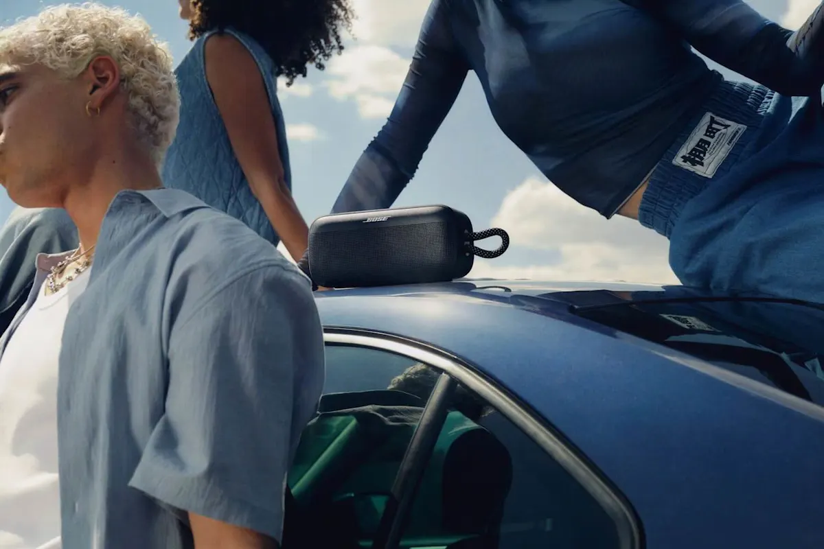 Ein Bose SoundLink Plus-Lautsprecher steht auf einem Autodach. Zwei Personen sitzen am Auto, eine steht davor.
