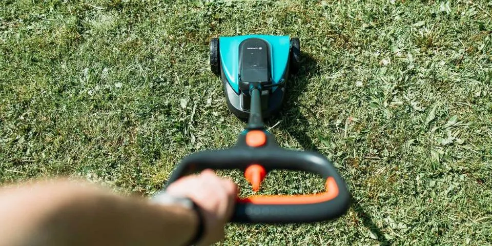 Person schiebt einen blauen Gardena Akku-Rasenmäher über eine Wiese und hält den schwarzen Griff mit orangefarbenen Elementen fest.