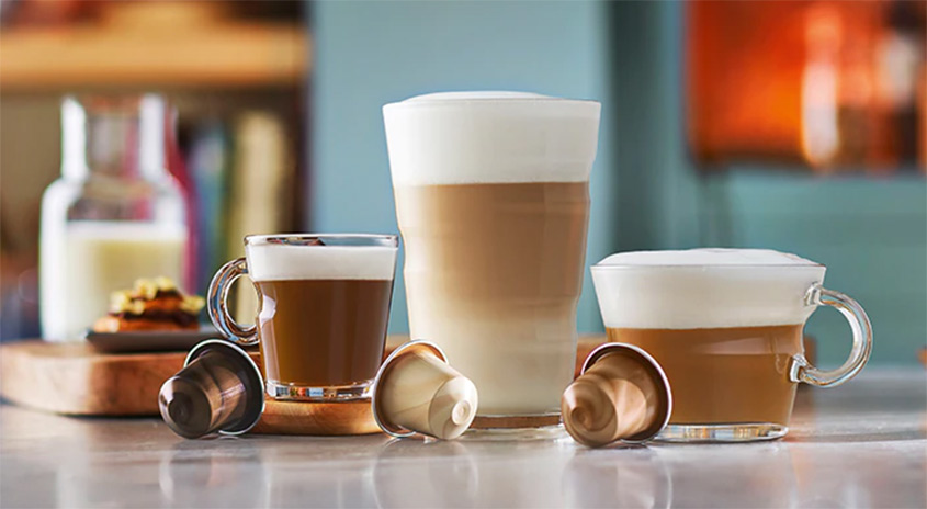 Drei Gläser mit Getränken, Kaffeekapseln im Vordergrund. Im Hintergrund eine Flasche Milch und ein Teller mit Speisen auf einem Brett.