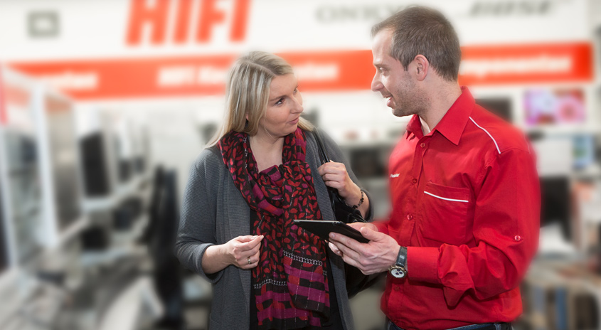 Ein Mann im roten Hemd zeigt einer Frau mit Schal ein Tablet in einem Geschäft. Im Hintergrund ist Werbung sichtbar.