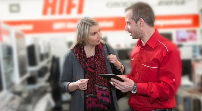 Ein Mann im roten Hemd zeigt einer Frau mit Schal ein Tablet in einem Geschäft. Im Hintergrund ist Werbung sichtbar.
