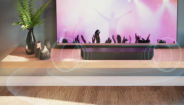 Auf einem Holzschrank stehen eine Soundbar, eine TV und Vasen. Der TV zeigt eine Menschenmenge mit erhobenen Händen.