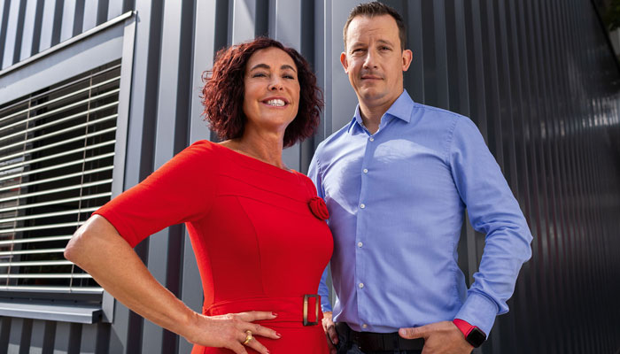 Ein Mann in blauem Hemd und eine Frau in rotem Kleid stehen vor einer Wand mit Fensterläden.