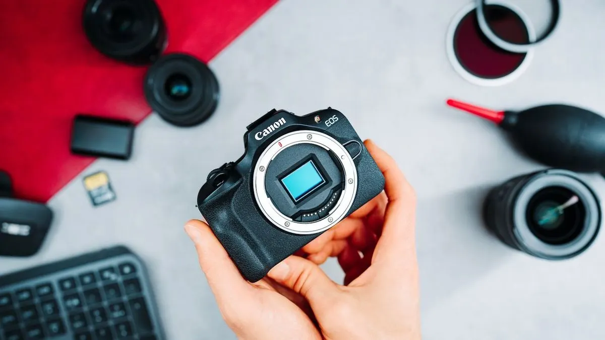 Hand hält schwarze Canon EOS Kamera mit blauem Sensor. Objektive, SD-Karte, Blasebalg, Filter, Tastatur auf rot/grauer Fläche.