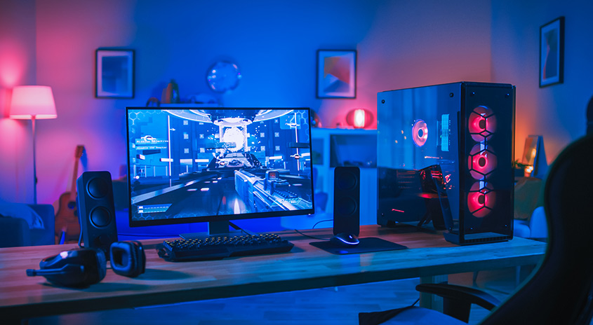 Ein Schreibtisch mit einem Computer, Lautsprechern, Kopfhörern und einer Tastatur. Der Computer hat rote Lüfter. Der Raum ist blau und pink beleuchtet.