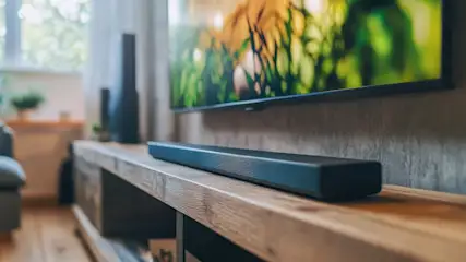 Schwarze Soundbar auf einem Holz-TV-Möbel, davor ein Fernseher mit grüner Landschaft, mit Betonwand und Fenster im Hintergrund.