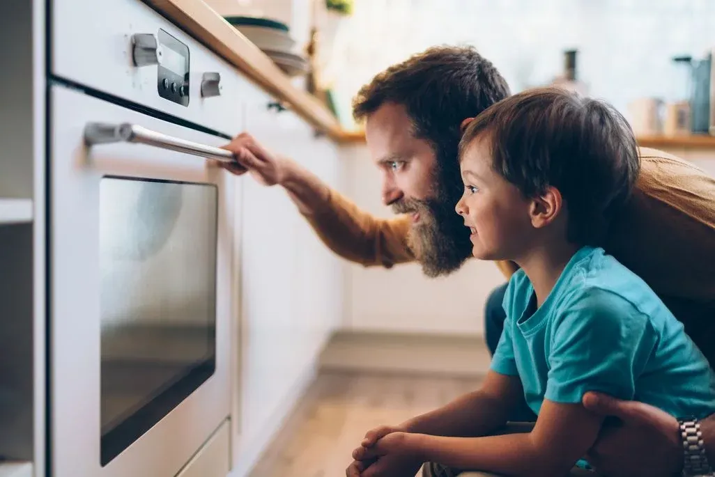 Vater und Kind öffnen gemeinsam Backofen in moderner Küche während Backvorgang.