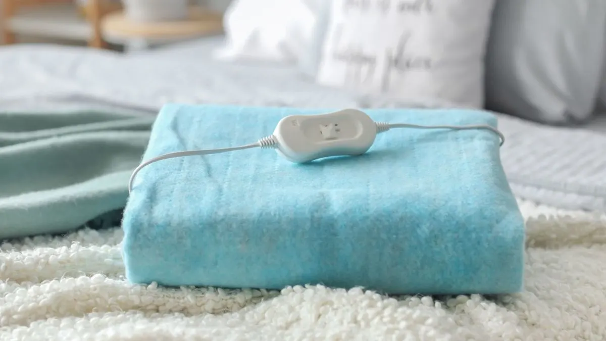 Zusammengefaltete blaue Wärmeunterlage mit weißem Kabel und Bedienelement liegt auf einem Bett.