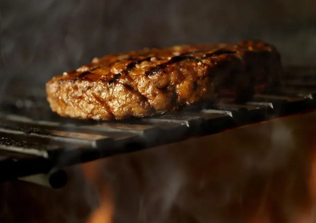 Ein dickes, braunes Fleischpatty mit Grillstreifen gart auf schwarzem Grillrost. Rauch steigt auf, darunter lodern orange Flammen.