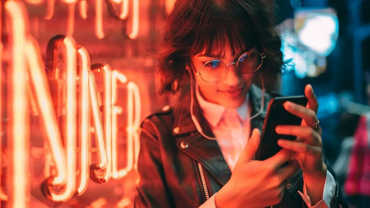 Person mit dunklen Haaren und Lederjacke steht vor einem leuchtenden Neon-Schriftzug und hält ein Smartphone in der Hand.