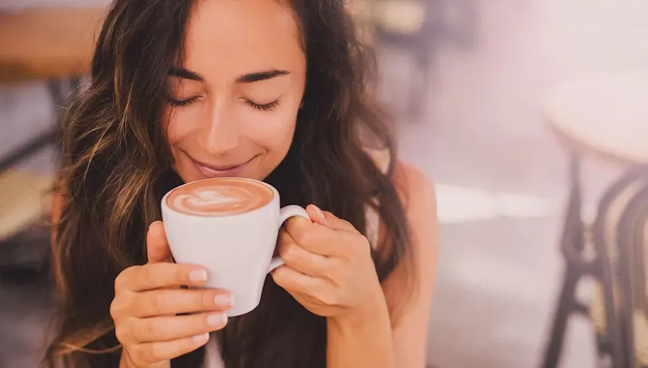 Eine lächelnde Person hält eine weiße Tasse mit Latte Art Kaffee und riecht daran. Im verschwommenen Hintergrund sind Tische und Stühle eines Cafés.