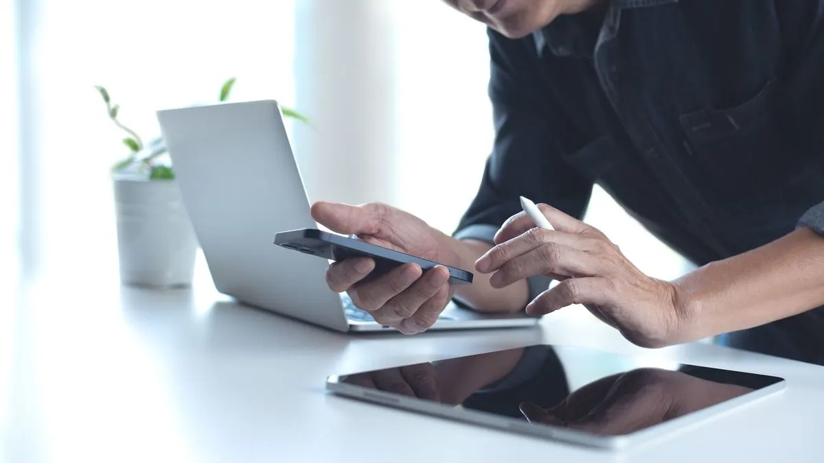 Person hält Smartphone und Stift, tippt auf Tablet. Laptop und Topfpflanze auf hellem Schreibtisch im Hintergrund.