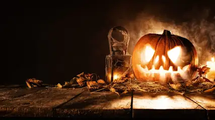 Eine gruselig beleuchtete Halloween-Kürbislaterne mit geschnitztem Gesicht steht auf einem alten Holztisch, umgeben von trockenem Laub, Spinnweben, einer alten Laterne und brennenden Kerzen im rauchigen Halbdunkel.