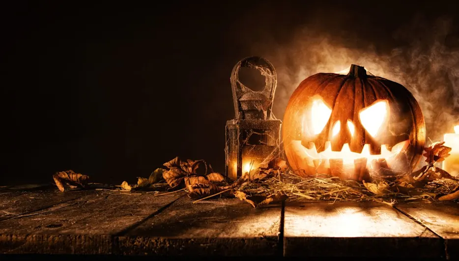 Eine gruselig beleuchtete Halloween-Kürbislaterne mit geschnitztem Gesicht steht auf einem alten Holztisch, umgeben von trockenem Laub, Spinnweben, einer alten Laterne und brennenden Kerzen im rauchigen Halbdunkel.