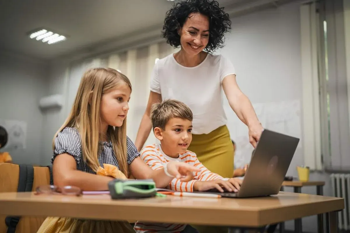 Lehrerin zeigt zwei Volksschülern etwas auf einem Laptop; beide sitzen am Tisch und schauen interessiert.