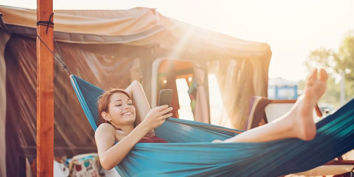 Eine lächelnde Person liegt in einer blauen Hängematte und schaut auf ein Smartphone. Im Hintergrund ist ein beiges Zelt und ein Holzpfosten bei Sonnenlicht zu sehen.