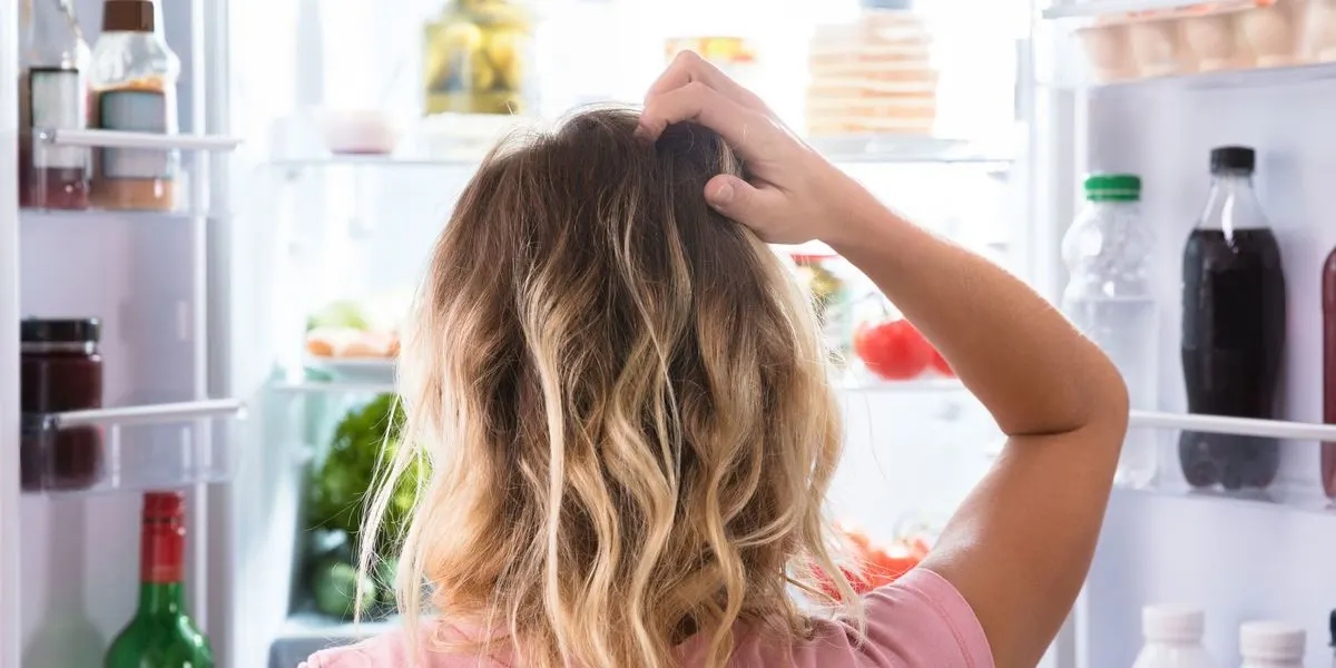 Person blickt in einen offenen, hellen Kühlschrank, kratzt sich am Kopf. Helle Regale voller Flaschen, Gläser und Lebensmittel.