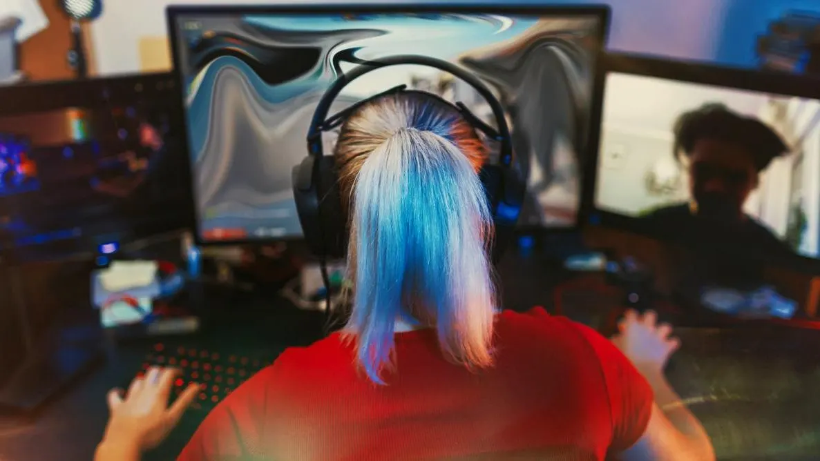 Person mit Headset sitzt in rotem T-Shirt an einem Schreibtisch vor mehreren Monitoren und spielt ein Computerspiel.