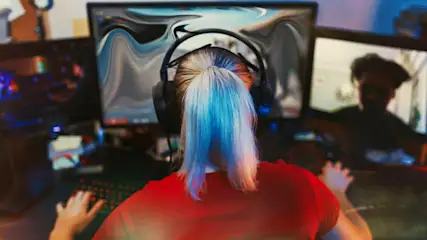 Person mit Headset sitzt in rotem T-Shirt an einem Schreibtisch vor mehreren Monitoren und spielt ein Computerspiel.