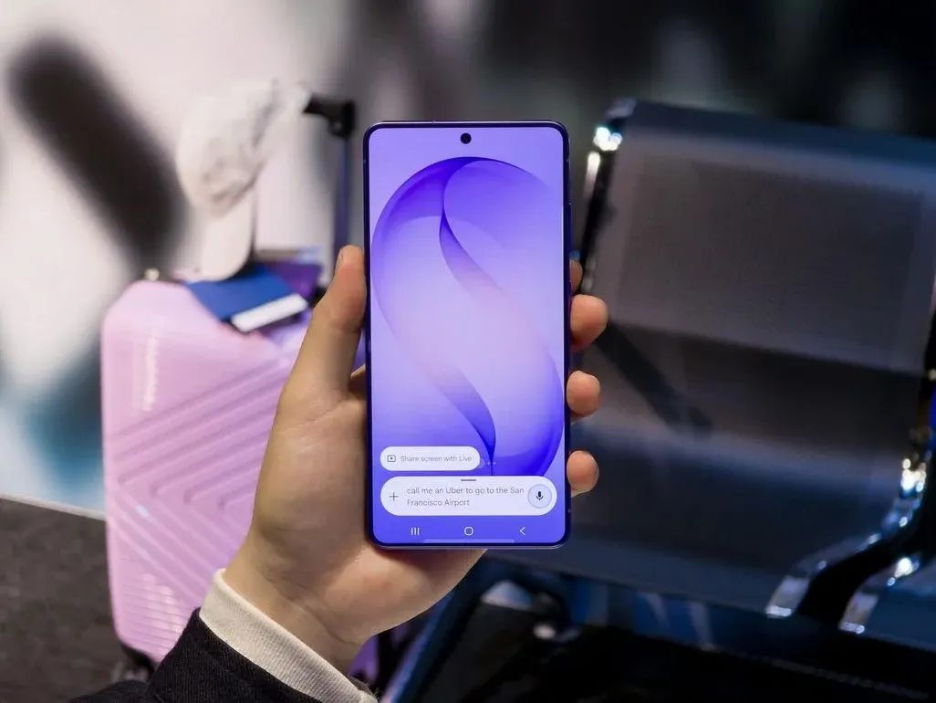 Hand hält ein modernes Smartphone mit großem Display in einem Wartebereich, im Hintergrund steht ein rosa Koffer neben Metall-Sitzbänken.