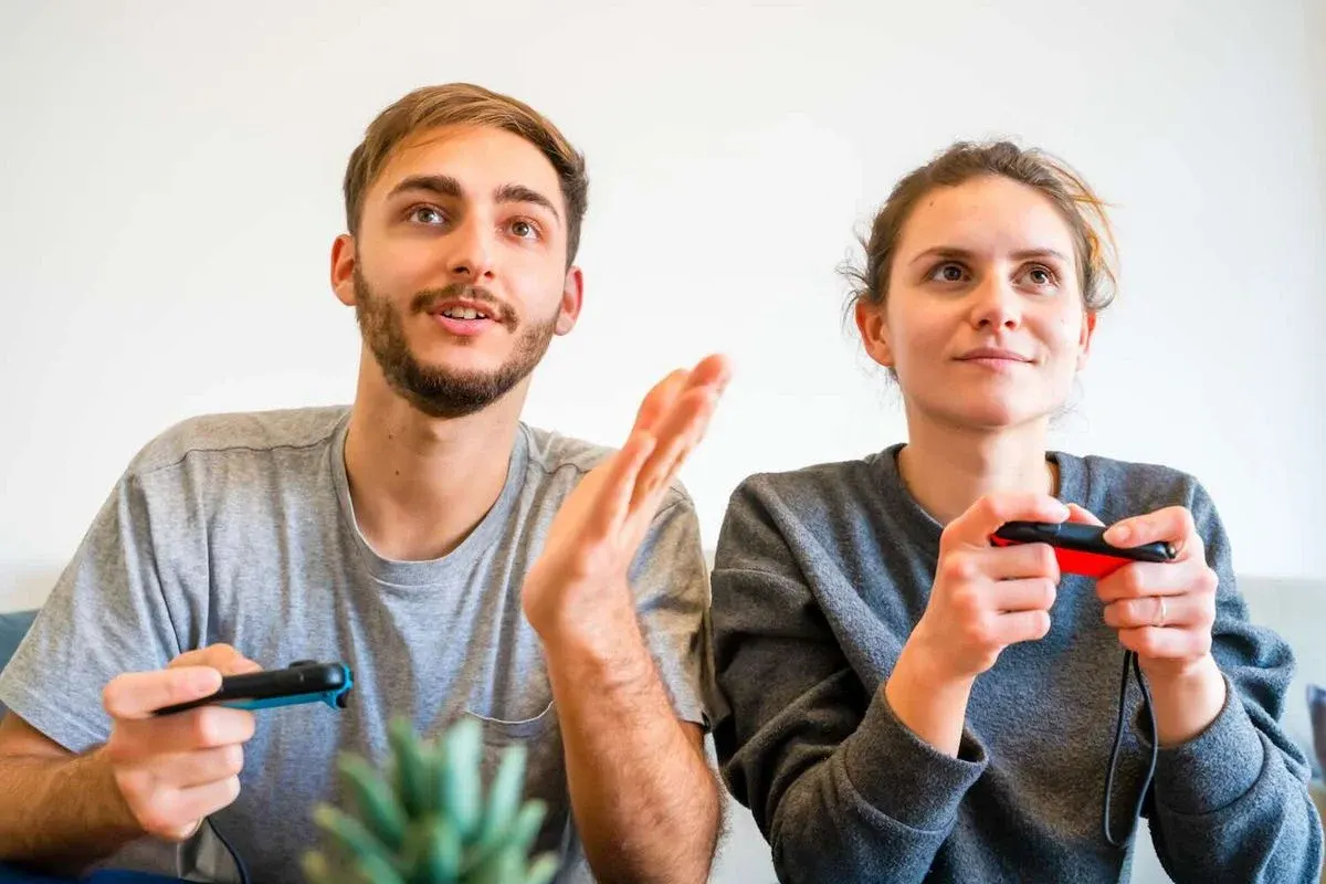 Zwei junge Erwachsene sitzen nebeneinander auf dem Sofa und spielen Videospiele; beide halten Nintendo Switch Joy‑Con‑Controller und schauen konzentriert auf den Bildschirm.