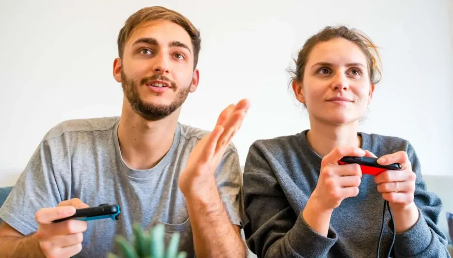 Zwei junge Erwachsene sitzen nebeneinander auf dem Sofa und spielen Videospiele; beide halten Nintendo Switch Joy‑Con‑Controller und schauen konzentriert auf den Bildschirm.