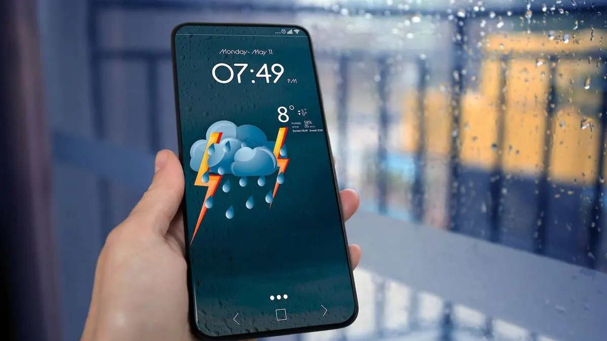 Hand haelt ein Smartphone mit Wetter-App, die Gewitter mit Regen und 8 Grad anzeigt, vor einem verregneten Fenster.