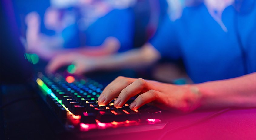 Nahaufnahme einer Hand, die auf einer schwarzen Tastatur mit farbiger Hintergrundbeleuchtung tippt, auf einem rosafarbenen Tisch.