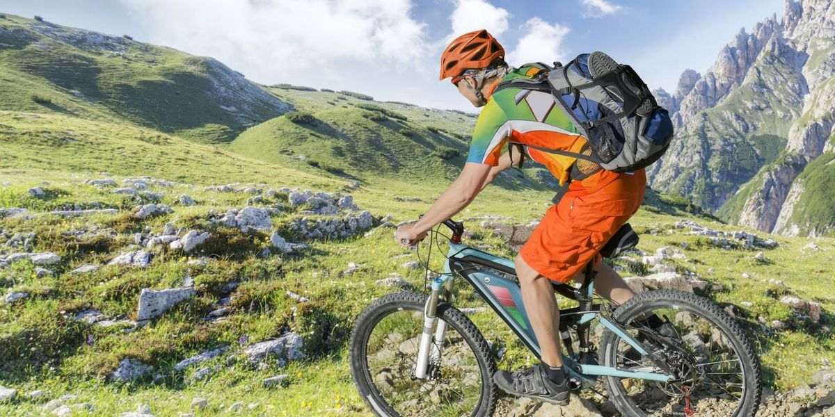 Radfahrer mit orangefarbenem Helm und Rucksack fährt auf einem hellblauen Mountainbike durch felsiges Grasland vor grünen Hügeln und grauen Bergen.