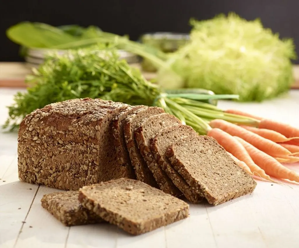 Geschnittenes Vollkornbrot mit Kernen liegt neben frischen Möhren und Gemüse.