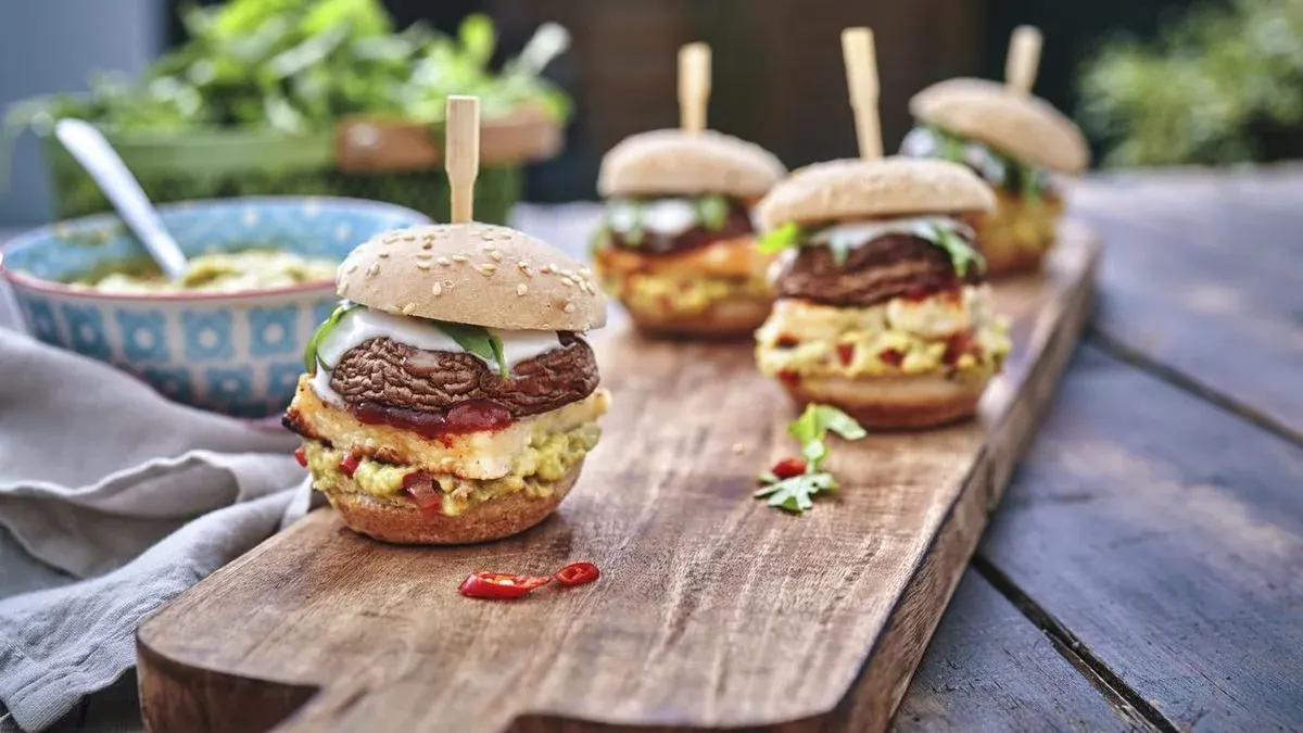Vier Mini-Burger mit Pilz-Patties, cremiger Soße, grünen Blättern und Guacamole auf Holzbrett. Dipschale und Salat im Hintergrund.
