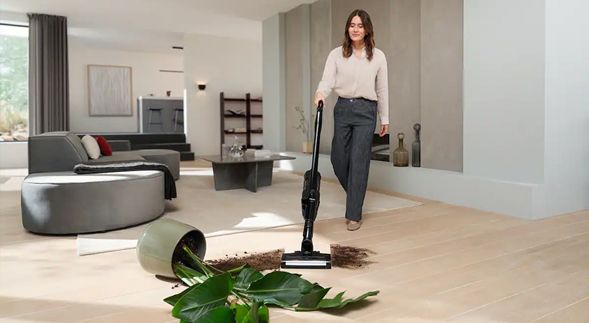 Person saugt mit dem Miele Triflex HX3 verschüttete Blumenerde einer umgekippten Topfpflanze von einem hellen Holzboden im modernen Wohnzimmer.