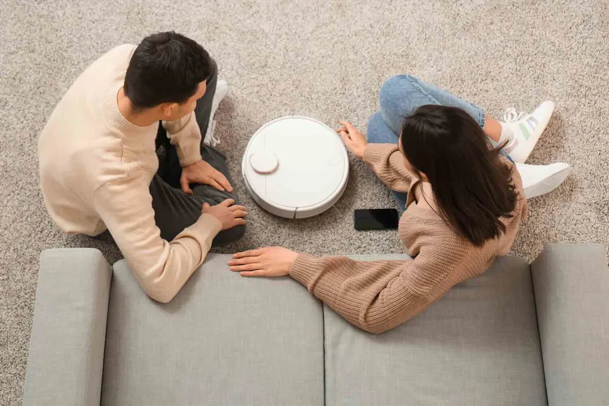 Ein Mann und eine Frau sitzen vor einer grauen Couch auf einem Teppichboden. Zwischen ihnen steht ein Saugroboter, daneben liegt ein Smartphone.