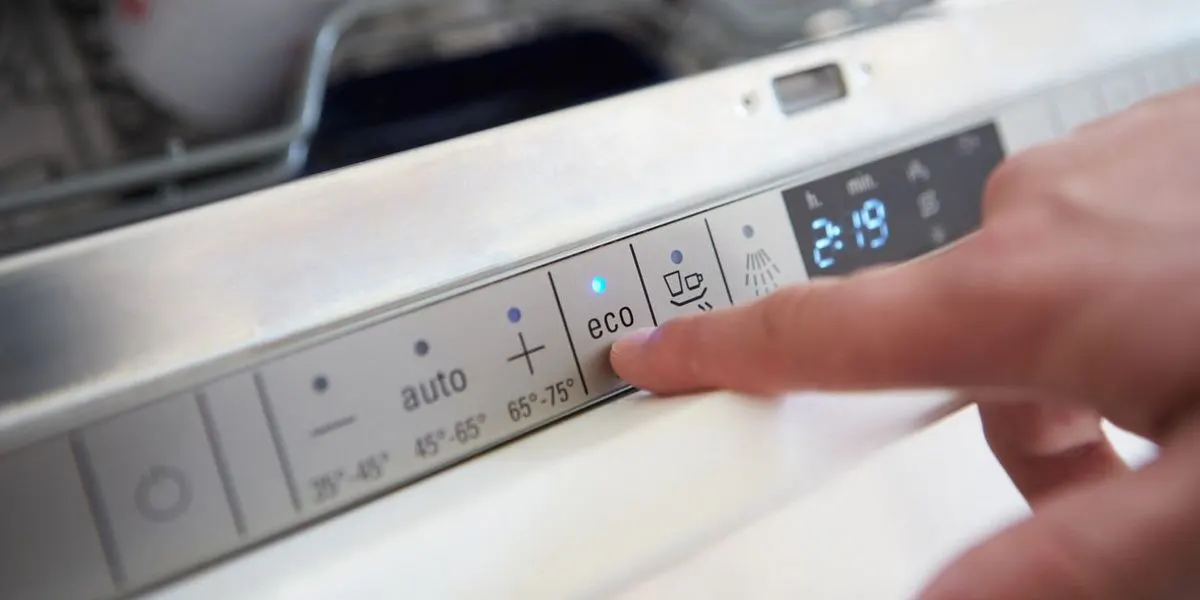 Finger drückt die "eco"-Taste auf einem Spülmaschinen-Bedienfeld mit blauer Anzeigeleuchte und der Zeit "2:19". Im Hintergrund sind Geschirrkörbe sichtbar.