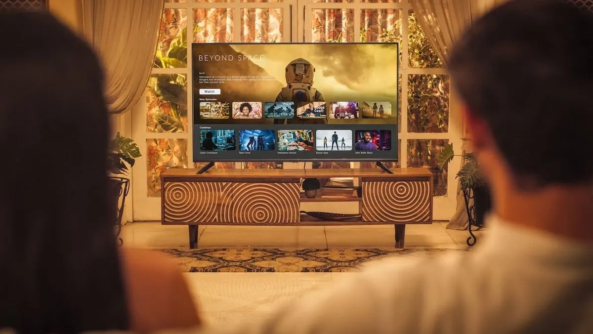 Zwei Personen blicken auf einen Fernseher mit Streaming-Interface auf einem Holzschrank mit Kreismustern. Hinten: Fenster und Pflanzen.