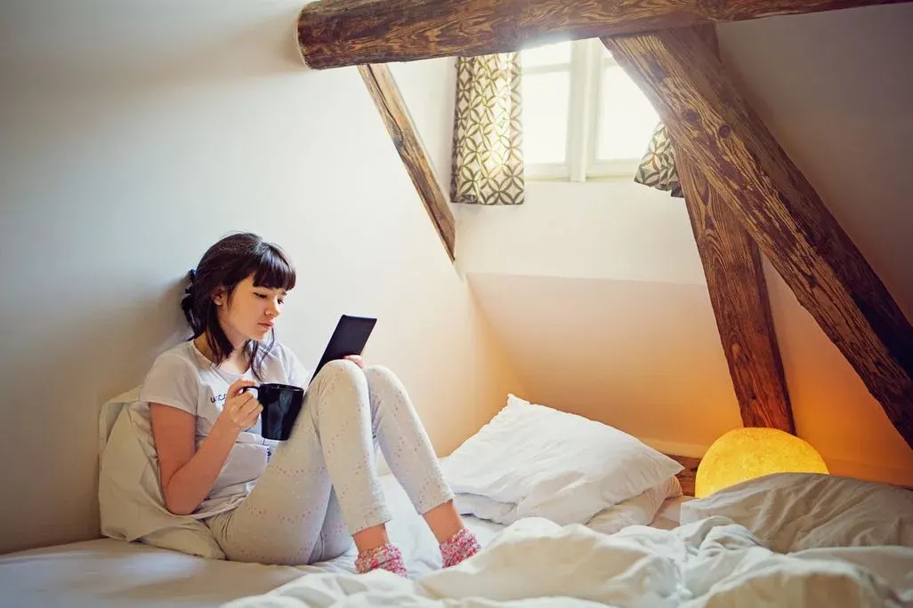 Eine Person sitzt im Bett, hält einen schwarzen Becher und ein Tablet. Weiße Laken, Dachschräge, Holzbalken, Fenster mit Vorhängen und eine gelbe Lampe.