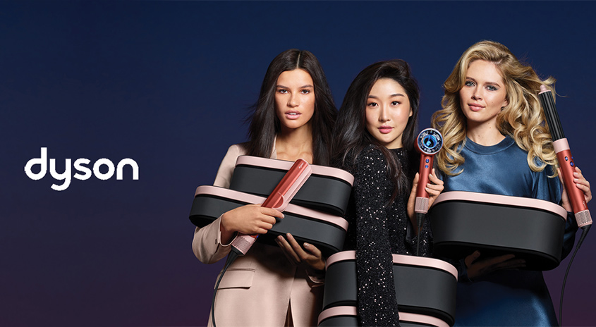Drei Personen halten Dyson-Haarstyling-Sets mit Werkzeugen in Schwarz und Roségold vor einem dunklen Hintergrund. "Dyson"-Logo links.