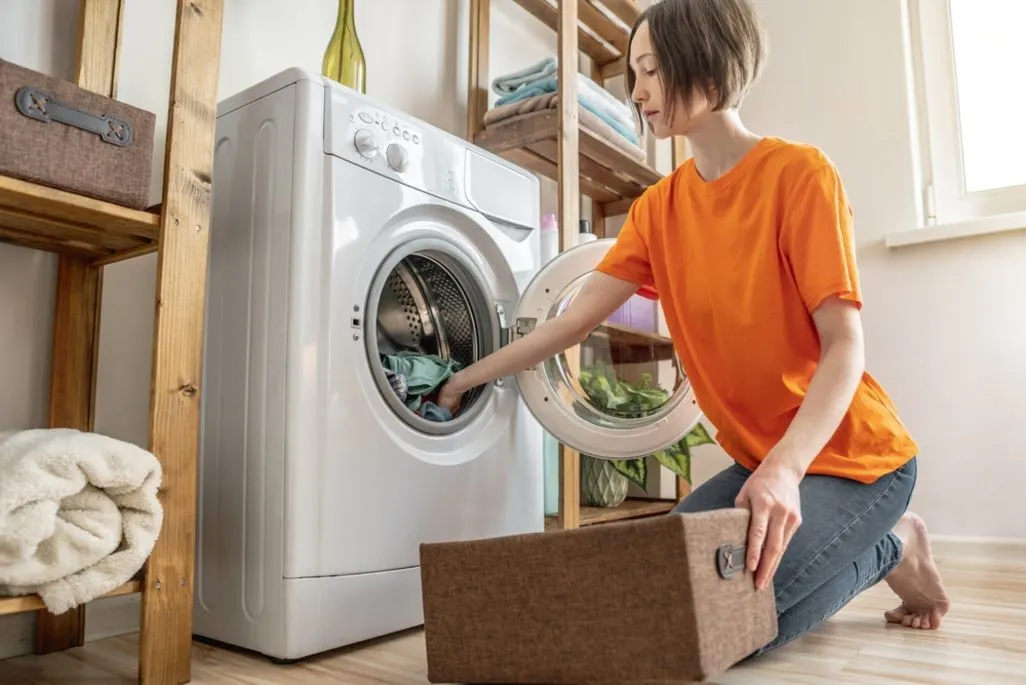 Frau in orangefarbenem Shirt kniet, belädt eine weiße Waschmaschine mit Wäsche. Braune Box am Boden. Holzregale mit Handtüchern im Hintergrund.