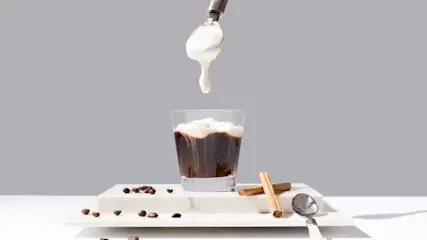 Schwarzer Kaffee in einem Glas auf den mit einem Löffel Milchschaum getropft wird, Zimtstangen und Kaffeebohnen als Deko daneben.